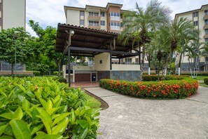 A covered outdoor area with a building in the background, surrounded by greenery and red flowers. - Costa Linda 8-11 3BR Apartment With Garden View (Jaco)