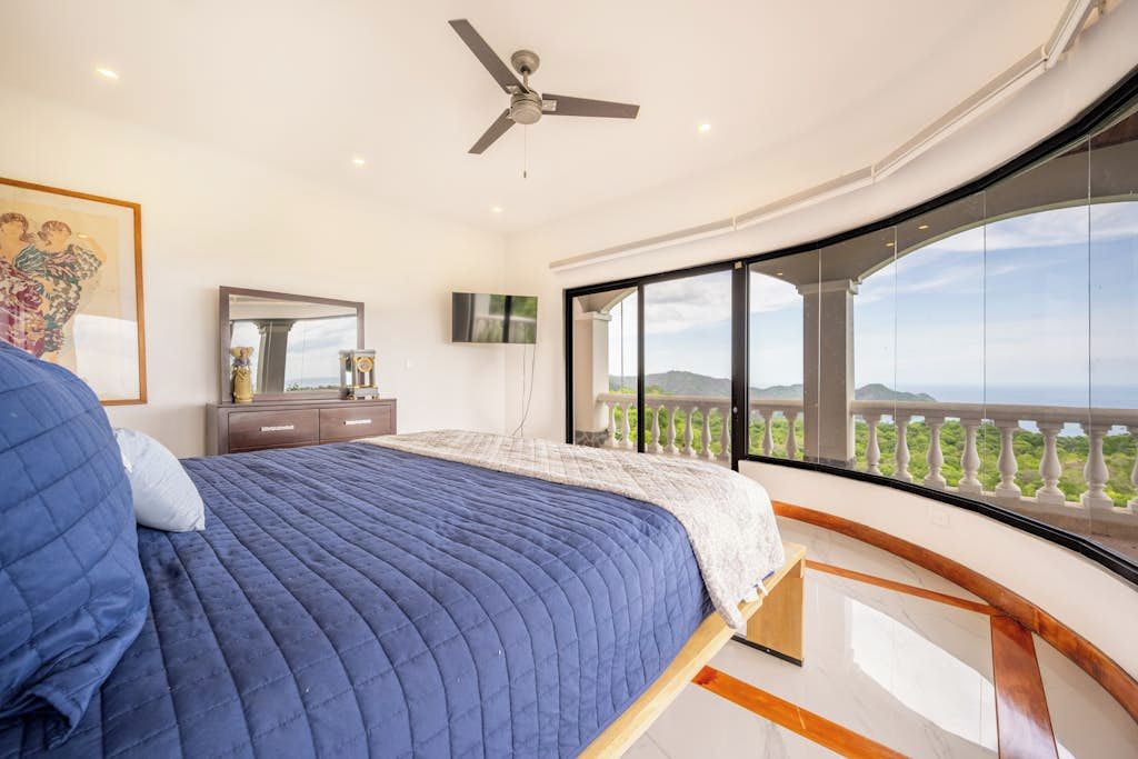 Une chambre à coucher avec un lit, un ventilateur de plafond, une télévision et une grande fenêtre donnant sur un paysage verdoyant.