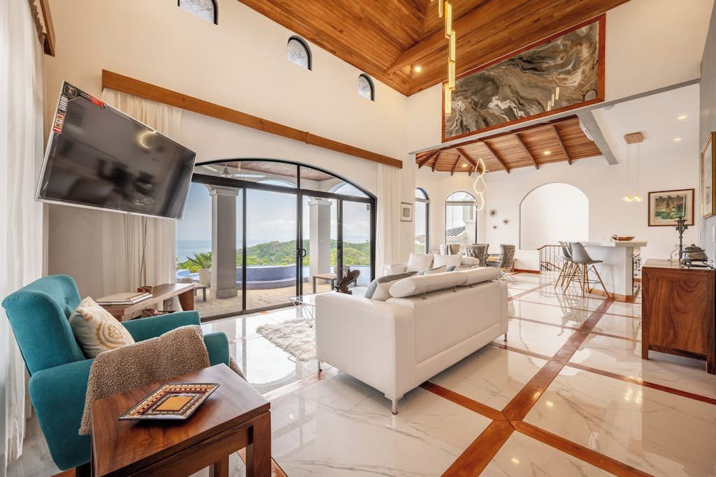 A spacious living area with a white sofa, teal armchair, and a wooden coffee table. A flat-screen TV is mounted on the wall, and there is a dining area with chairs and a table.