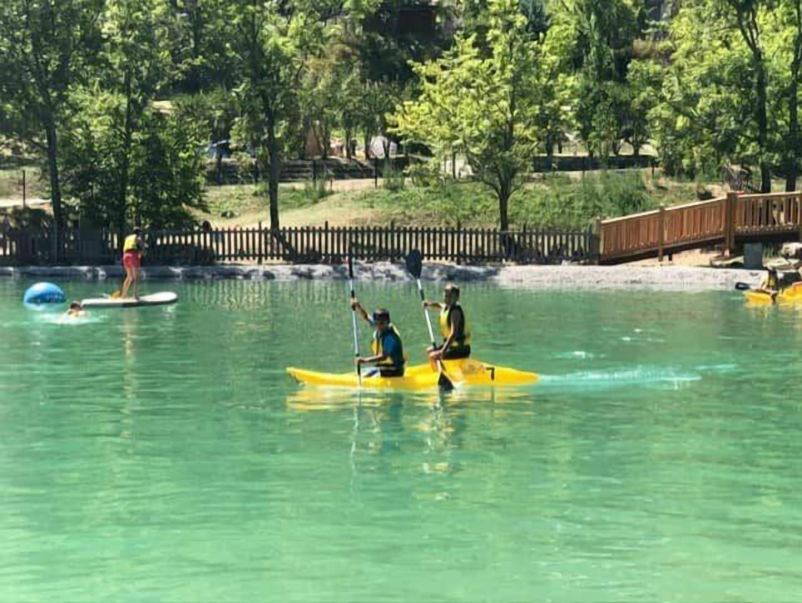 A kayaker and a paddleboarder on a calm body of water with trees and a wooden bridge in the background.