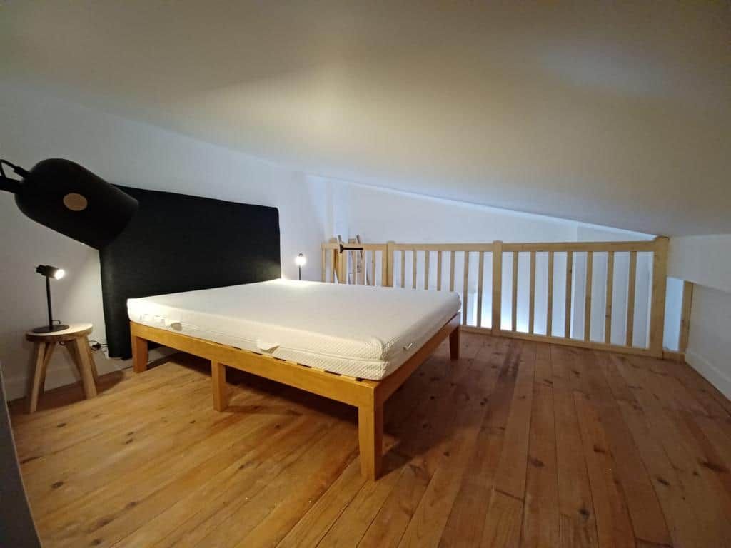 Un dormitorio con cama de madera, un taburete pequeño y una mesita de noche con lámpara.