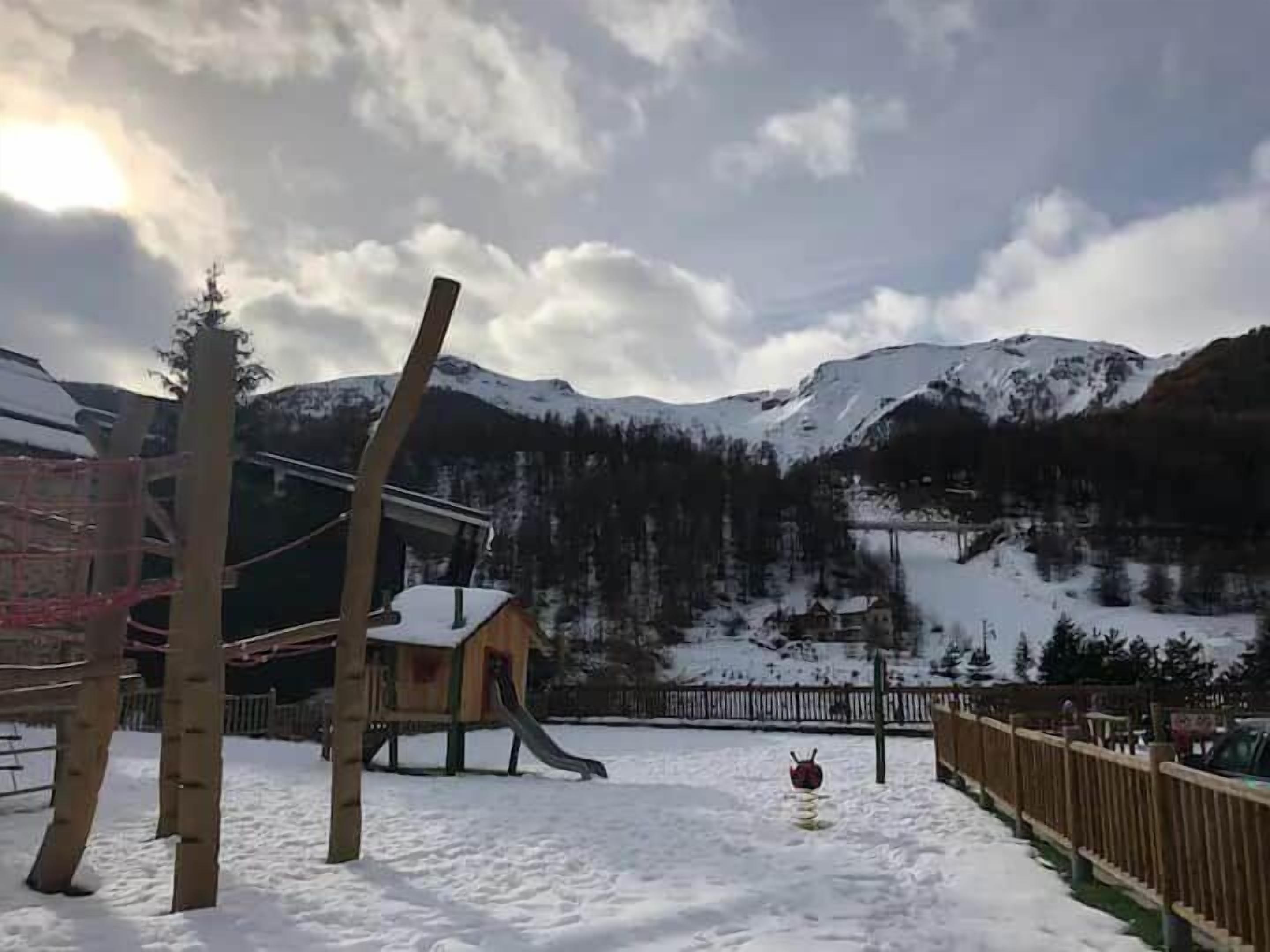 Un espacio exterior nevado con un parque infantil, una estructura de madera y vistas a montañas cubiertas de nieve.