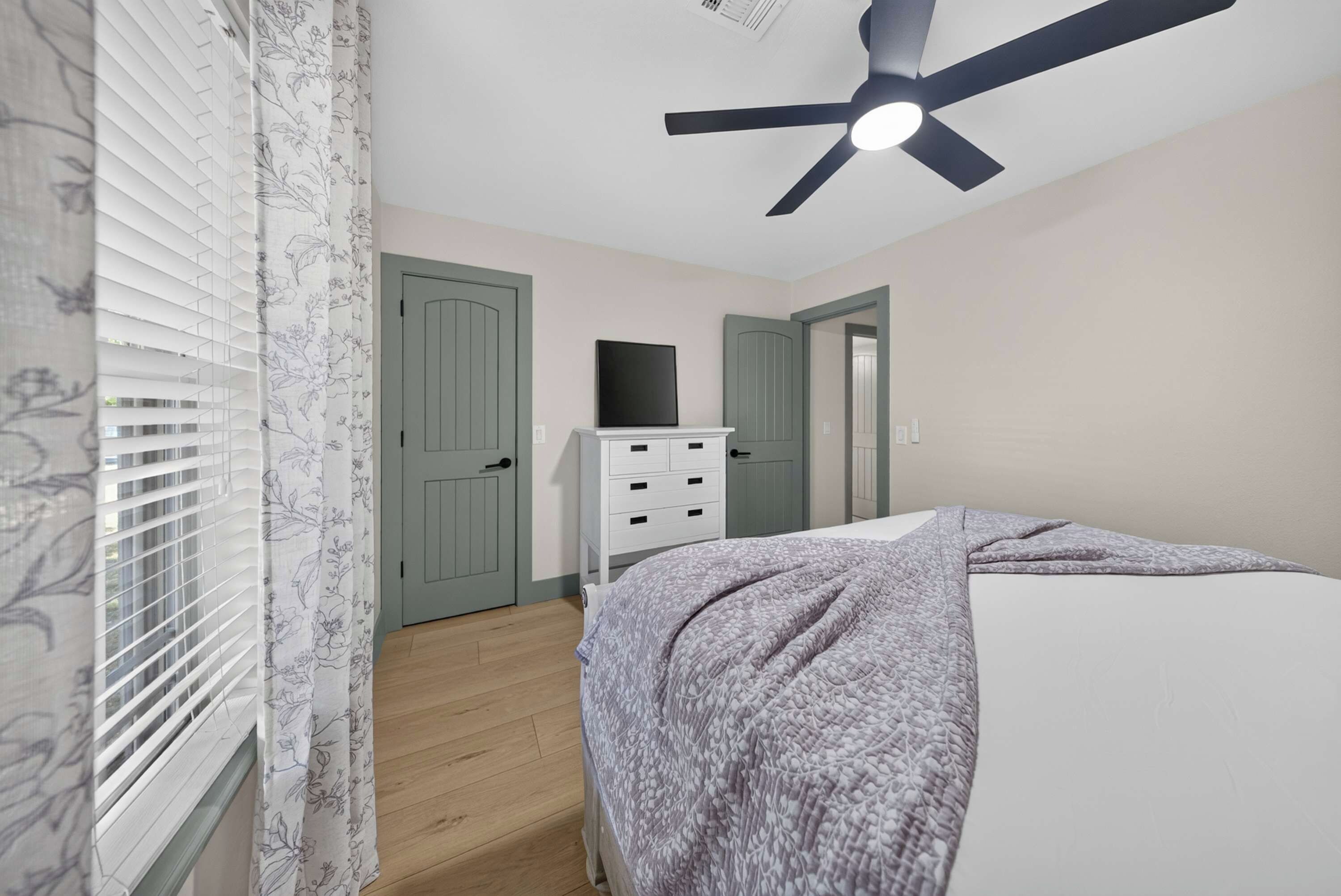 A bedroom with a bed, a dresser, a ceiling fan, and a door.