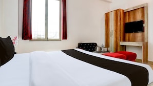 A hotel room with a bed, a TV mounted on a wooden panel, a window with curtains, and a small table with a kettle and glasses. - Deluxe Hotel Rk Heights And Banquets (Varanasi)