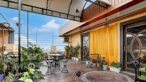 An outdoor patio with tables and chairs, surrounded by potted plants and a yellow curtain. - Deluxe Hotel Rk Heights And Banquets (Varanasi)