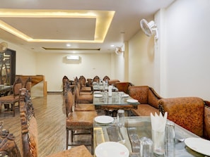 A restaurant with a long table set for dining, upholstered chairs, and a ceiling fan. - Deluxe Hotel Rk Heights And Banquets (Varanasi)