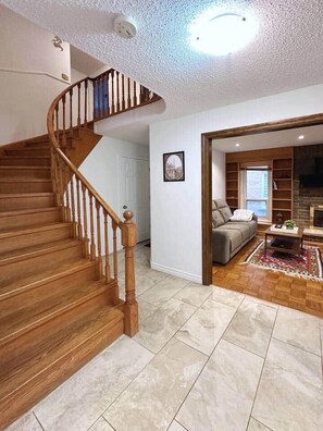 Eine Treppe mit Holzspindeln führt zu einer Empore, ein Wohnzimmer mit Sofa und Kamin, und ein Blick ins Freie durch ein Fenster.