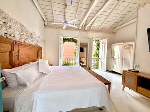 Une chambre à coucher avec une tête de lit en bois, un grand lit avec une literie blanche, un ventilateur de plafond et une vue sur le balcon extérieur.