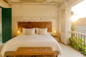 Een slaapkamer met een groot bed, twee nachtkastjes met lampen, een houten bankje, een groene deur en een balkon met planten.