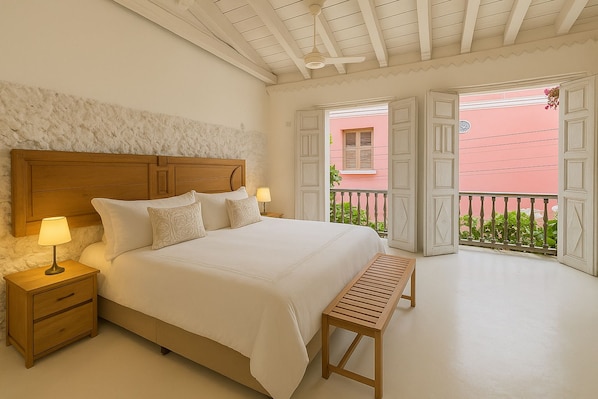 Een slaapkamer met een houten hoofdeinde, een bed met witte lakens, twee bedlampen, een houten bankje en een balkon met open deuren.