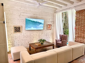 A living room with a white brick wall, a flat-screen TV, a wooden coffee table, a beige sofa, and a patterned armchair. - by Calamari.Homes - Casa Aura Victoria - Visitors permitted (Cartagena de Indias)