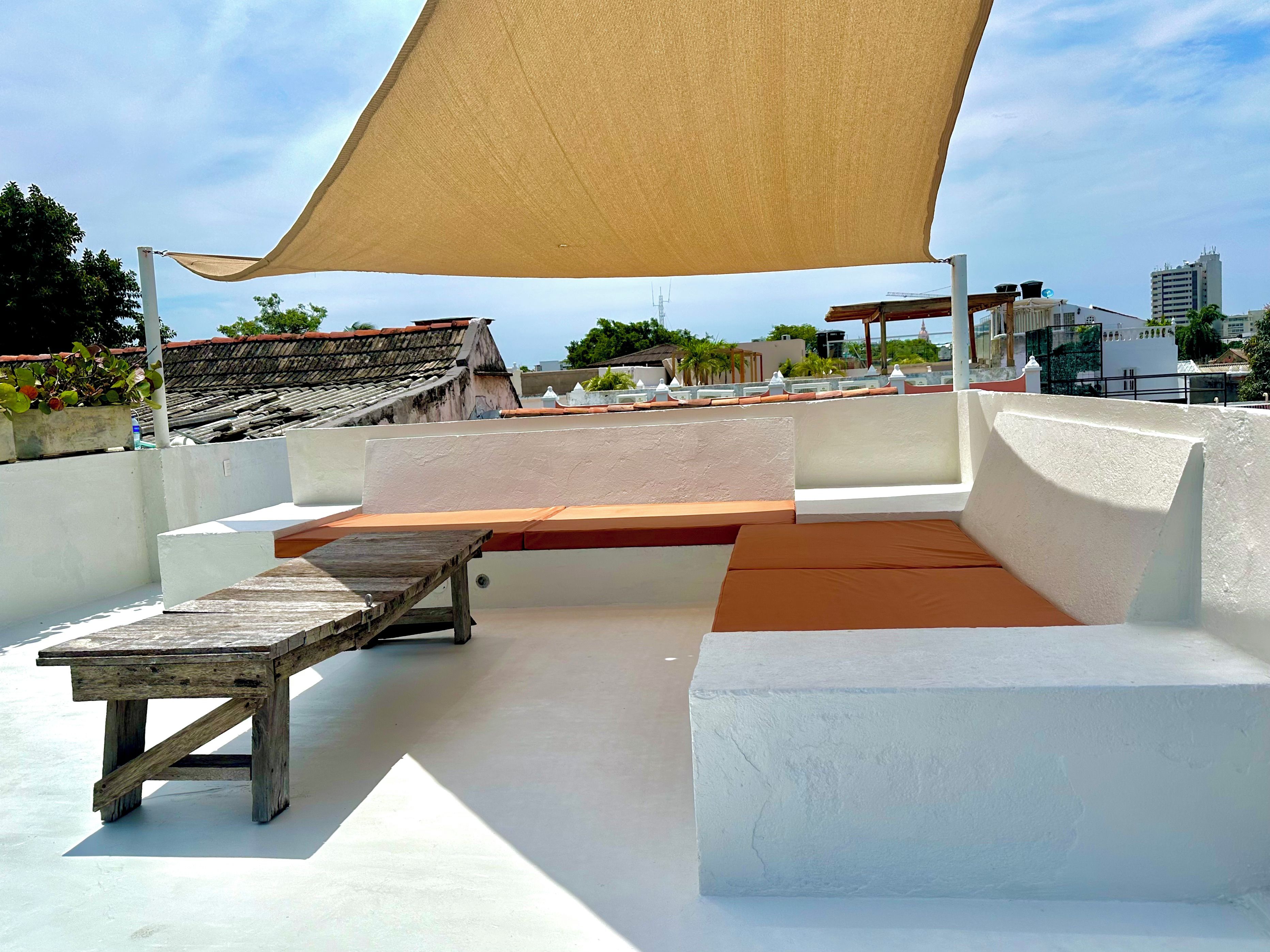 A rooftop terrace with a wooden table, a cushioned bench, and a large shade sail.