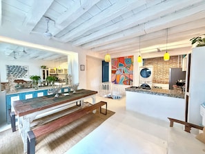A modern kitchen with a central island, a dining area with a wooden bench, and a colorful abstract painting on the wall. - by Calamari.Homes - Casa Aura Victoria - Visitors permitted (Cartagena de Indias)