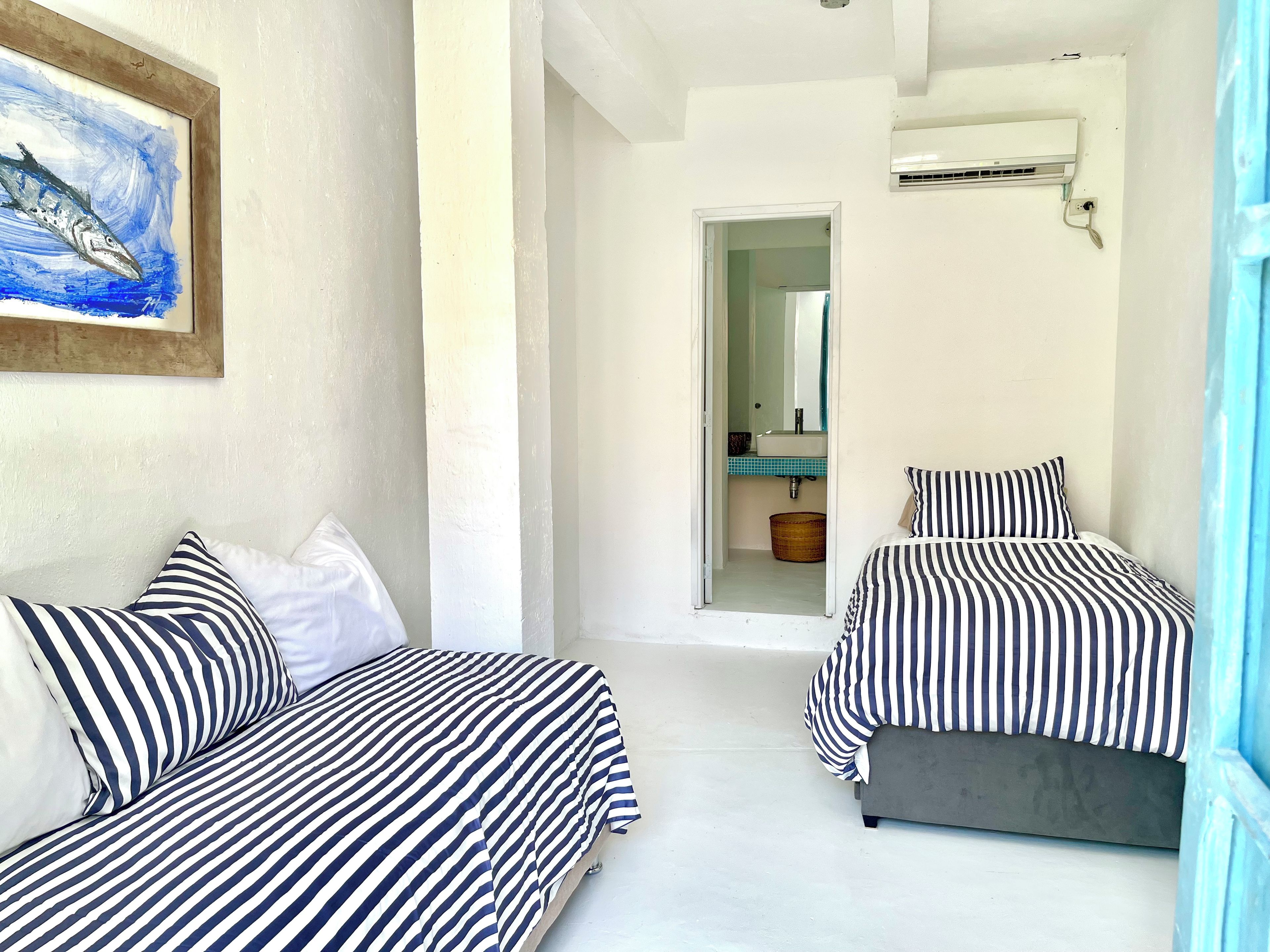 Quarto com duas camas, um quadro, banheiro visível por uma porta aberta e um ar condicionado.