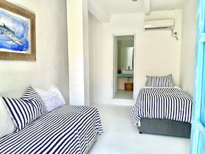 A room with two beds, a painting, a bathroom visible through an open door, and an air conditioning unit. - by Calamari.Homes - Casa Aura Victoria - Visitors permitted (Cartagena de Indias)