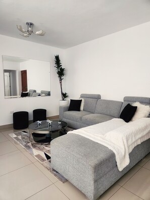 A modern living room with a grey sofa, a glass coffee table, a black ottoman, a large mirror, and a ceiling fan. - Fully Furnished Apartment in Miraflores (Miraflores)