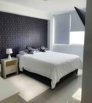 A modern bedroom with a large bed, a nightstand, a lamp, and a window with blinds. - Fully Furnished Apartment in Miraflores (Miraflores)
