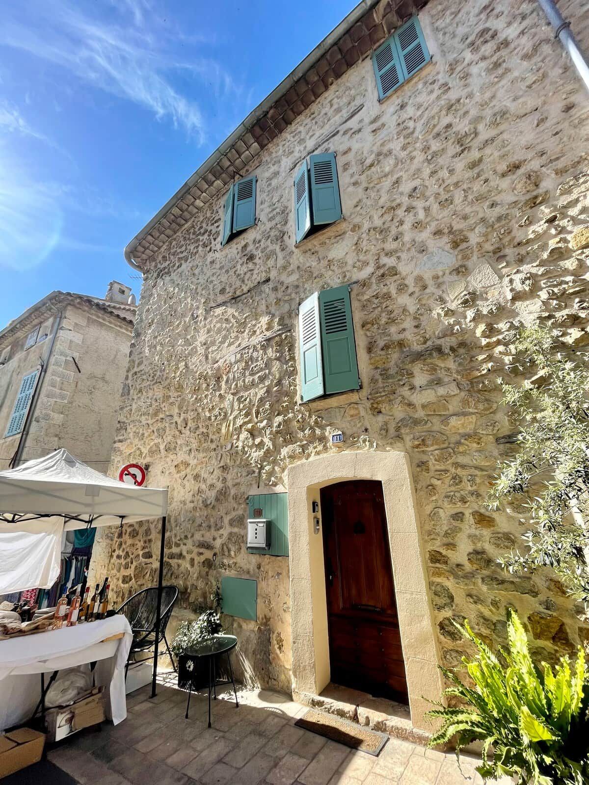 A stone building with a wooden door and shutters, a small outdoor seating area with a table and chairs, and a tent.