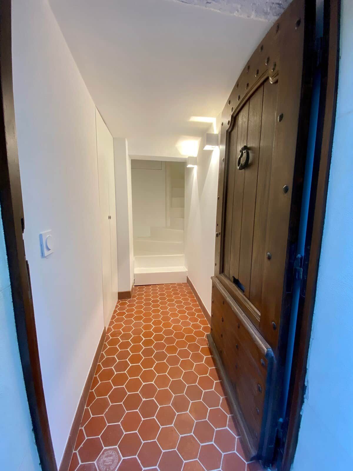 Un pasillo estrecho con suelo de baldosas, una puerta de madera y una escalera blanca al fondo.