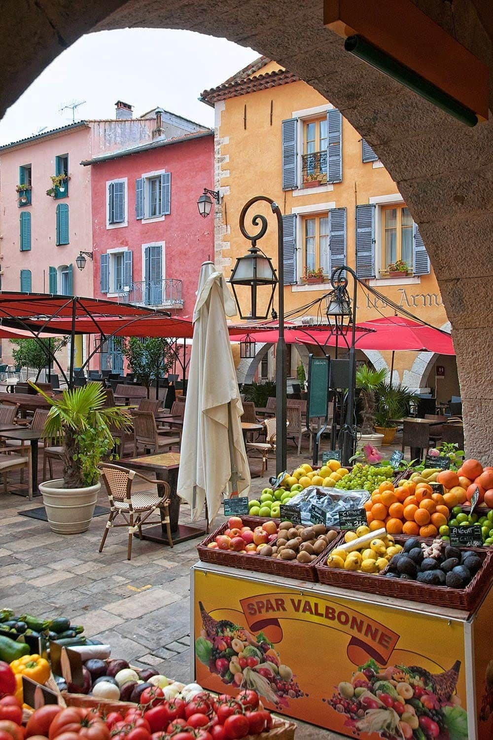An outdoor market with a fruit stand labeled 'SPAR VALBONNE' and a red umbrella.