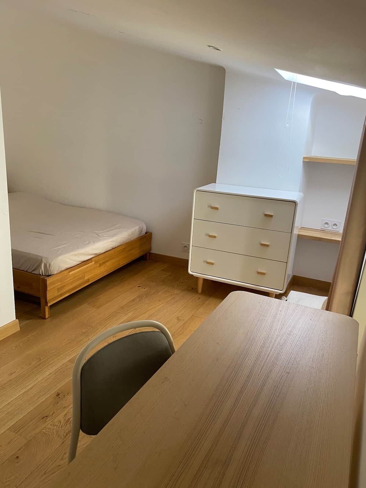 A small, sparsely furnished room with a wooden floor, a single bed, a white dresser, and a desk.