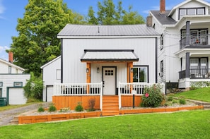 A two-story house with a white exterior, a front porch with a white picket fence, and a small garden area with plants and a flower bed. - Oswego River View Work Hub Near Phoenix (Phoenix)