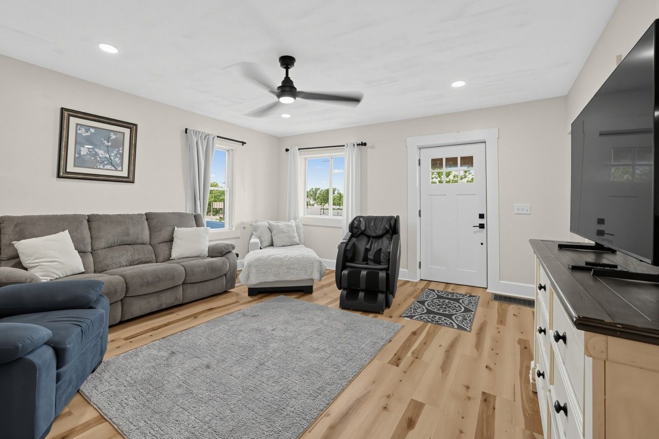 Un salon moderne avec un canapé gris, un canapé bleu, un fauteuil massant, un téléviseur à écran plat et un ventilateur de plafond.