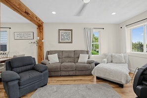 A living room with a grey sofa, a blue armchair, and a white bed. There is a framed picture on the wall and a window with curtains. - Oswego River View Work Hub Near Phoenix (Phoenix)