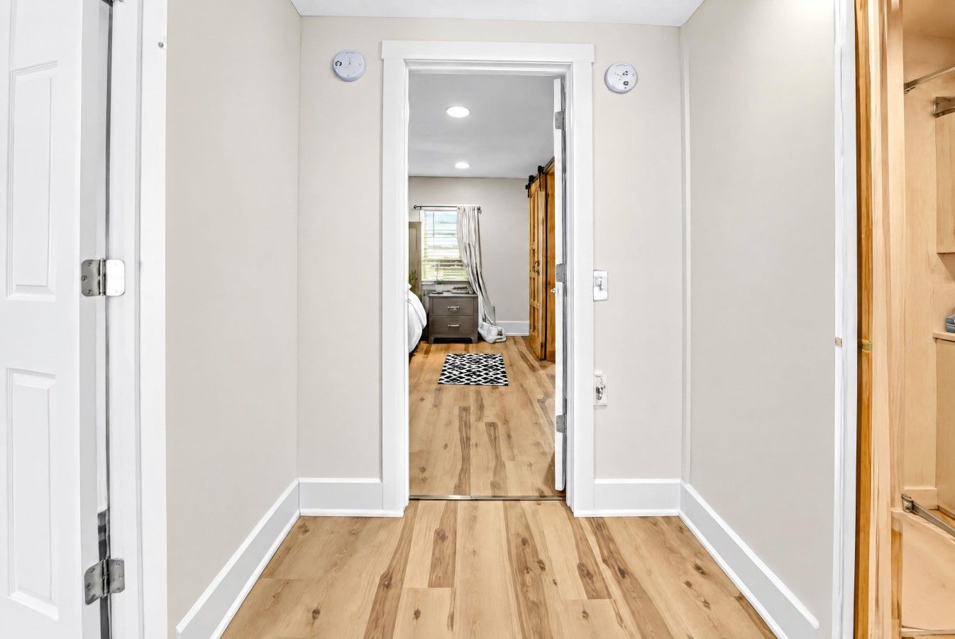 A hallway with a wooden floor leading to a room with a bed and a window.