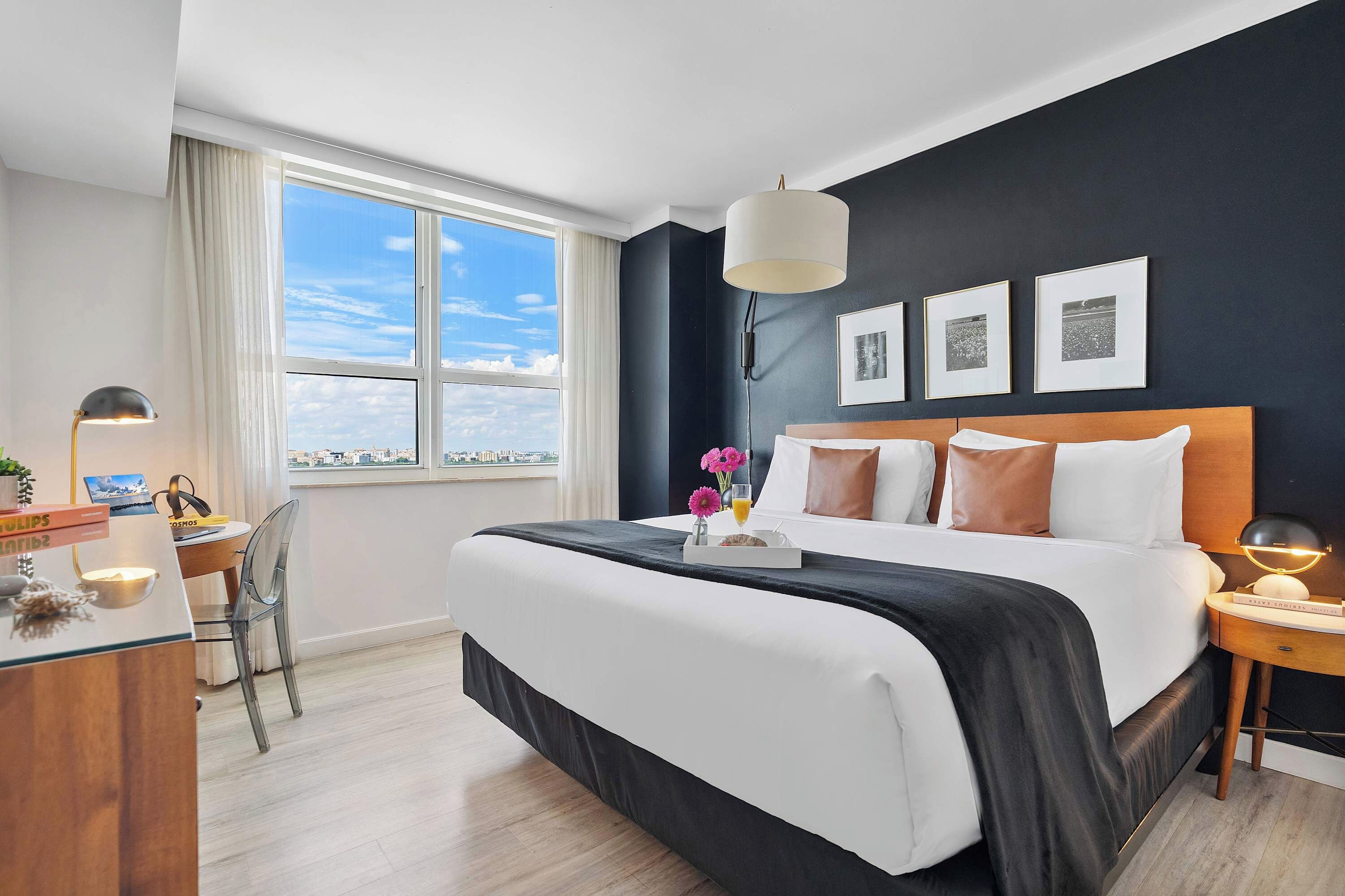 A hotel room with a large bed, a desk with a lamp, a chair, and a view of the city through the window.
