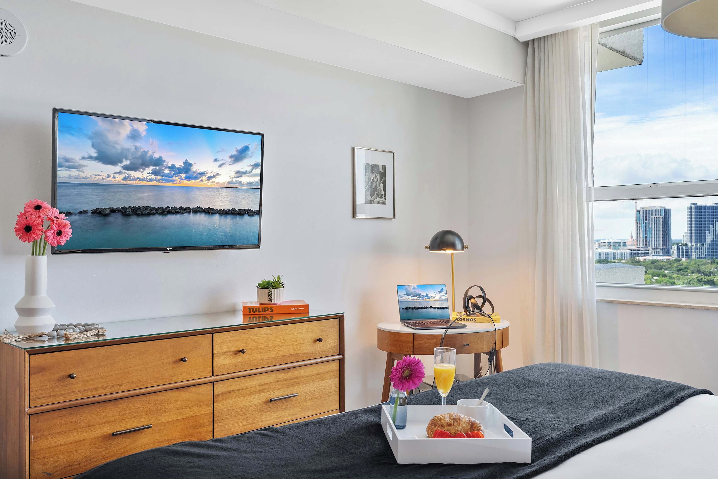 Habitación de hotel con cama, escritorio con una laptop, televisor con vista panorámica, ventana con vista a la ciudad y bandeja con artículos de desayuno.