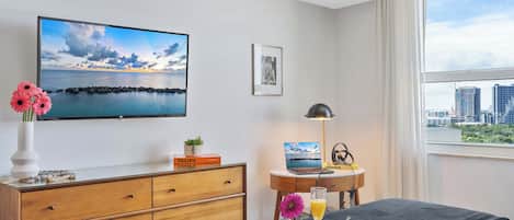 A hotel room with a bed, a desk with a laptop, a TV with a scenic view, a window with a cityscape view, and a tray with breakfast items.