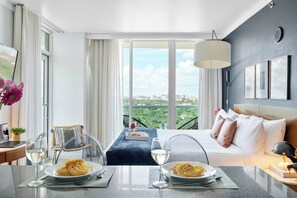 A hotel room with a bed, a dining table with plates of spaghetti, a laptop, a potted plant, and a view of the city. - Stylish Studio – Coconut Grove Retreat (Miami)