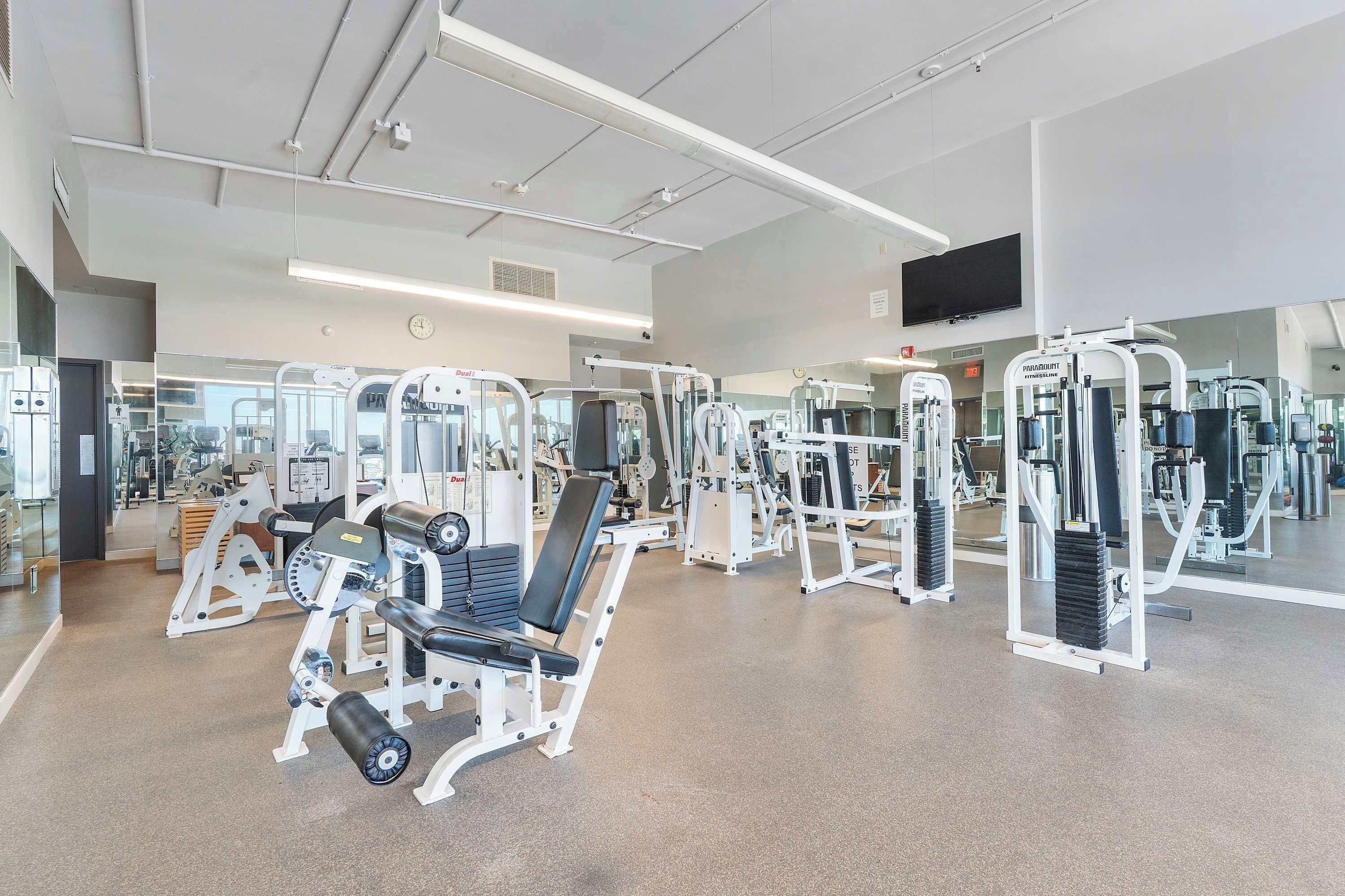 A modern gym with various exercise machines, mirrors, and a large TV screen.