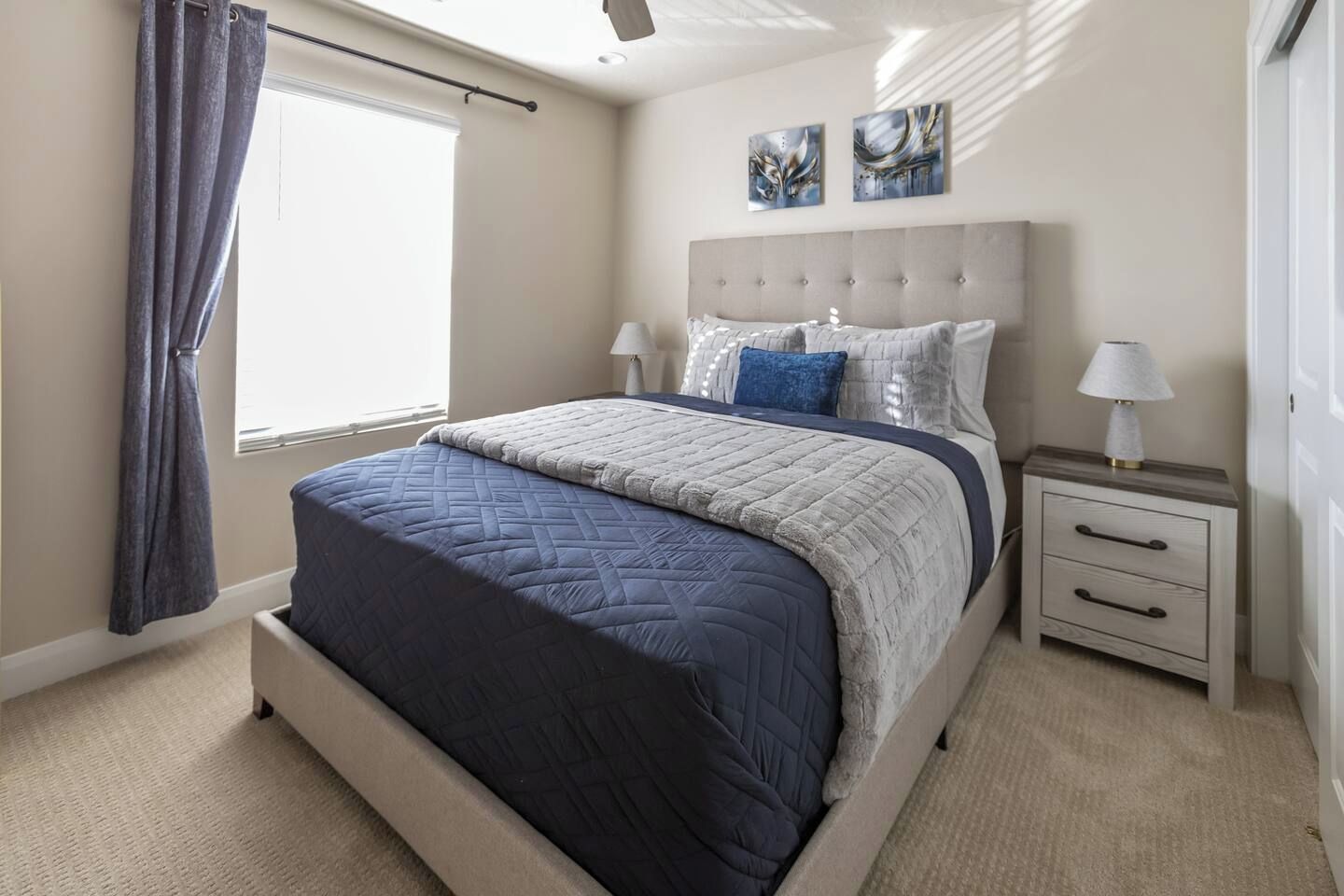 Un dormitorio con cama, mesitas de noche, una ventana con cortinas y una obra de arte en la pared.