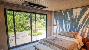 Un dormitorio con una cama grande, un banco, un cuadro en la pared y vistas a la vegetación a través de una puerta corrediza.