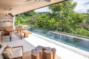 Een modern buitenzwembad met een houten terras, zitplaatsen en uitzicht op weelderige begroeiing.