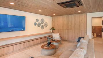 Ein modernes Wohnzimmer mit einem großen Flachbildfernseher an der Wand, einer Holzbalkendecke, einer Sitzecke mit Sofa und rundem Couchtisch sowie einem dekorativen Wandbild.