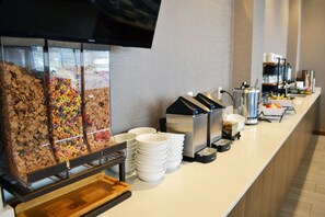A buffet area with cereal options, a coffee machine, and stacked white plates. - King 1BR Suite w/ Kitchen, Hot Breakfast & Pool (Red Deer)