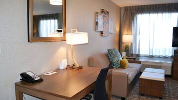 Habitación de hotel con un escritorio de madera, una silla, un sofá, una mesita, un televisor y una ventana con cortinas.