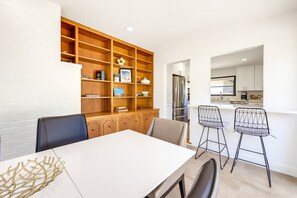 A modern kitchen with stainless steel appliances, a dining area with a white table and two bar-style chairs, and a wooden bookshelf filled with books and decorative items. - The Shores House,  Walk to Beach & Fire Pit (San Diego)