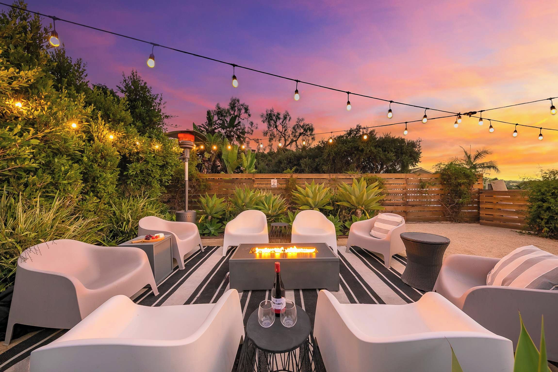 An outdoor seating area with white chairs and tables, a fire feature, and string lights against a sunset sky.