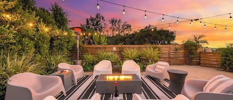 Un espacio al aire libre con sillas y mesas blancas, una chimenea y luces colgantes contra un cielo de atardecer.
