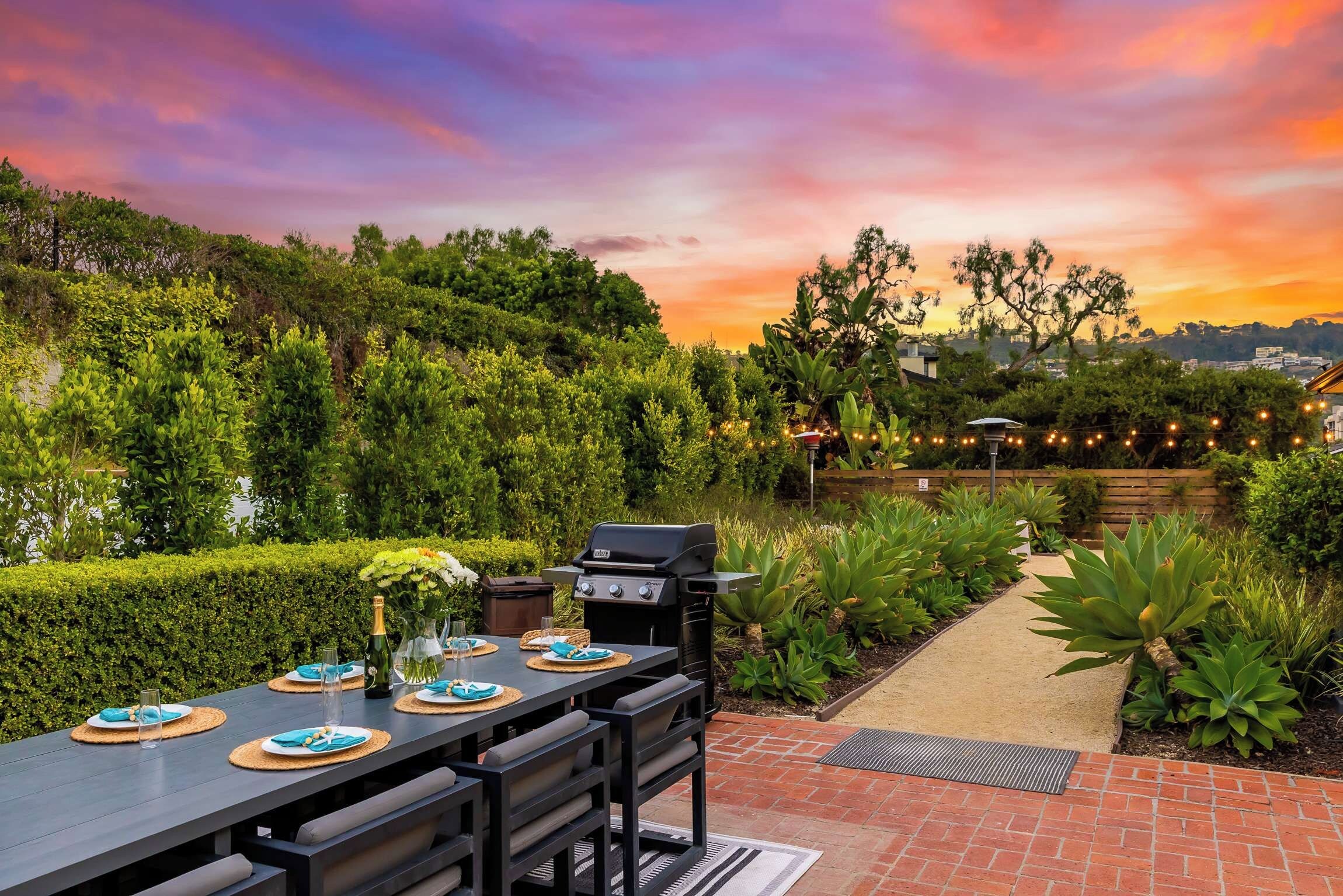 An outdoor dining area with a table set for four, a grill, and a view of a sunset.