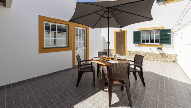 Een terras met een tafel, stoelen, een parasol en een raam met luiken.