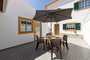 Une terrasse avec une table et des chaises, un parasol et une fenêtre avec des volets.