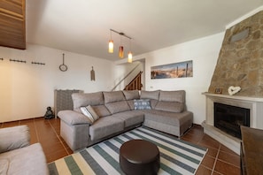 A living room with a sectional sofa, a fireplace, a staircase, and a wall-mounted clock. - Casa Onda Perfeita - Sunny Terrace (Peniche)