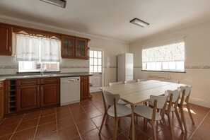 A kitchen with wooden cabinets, a built-in dishwasher, a dining table with chairs, and a window with patterned curtains. - Casa Onda Perfeita - Sunny Terrace (Peniche)