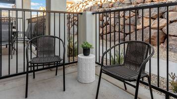 Two wicker chairs with a small potted plant on a white pedestal, set against a stone wall and metal railing.