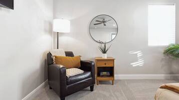 A room with a leather chair, a small wooden nightstand with a lamp, a round mirror, and a potted plant.